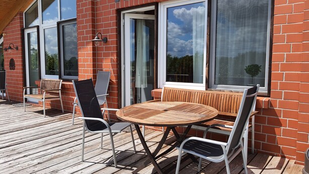 A wooden deck with a round table and chairs, a brick wall, and large windows. - Sund (Grosefehn)