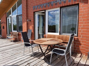 A wooden deck with a round table and chairs, a brick wall, and large windows. - Sund (Grosefehn)