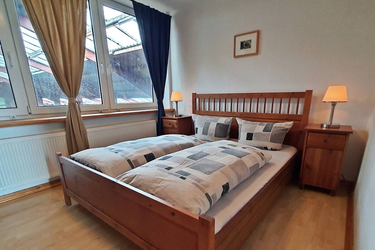 A double bed with a wooden headboard, two bedside tables with lamps, a window with curtains, and a framed picture on the wall.