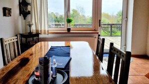 A dining area with a wooden table, chairs, a small table with a lamp, and a view of a balcony with a railing and greenery outside. - Sund (Grosefehn)