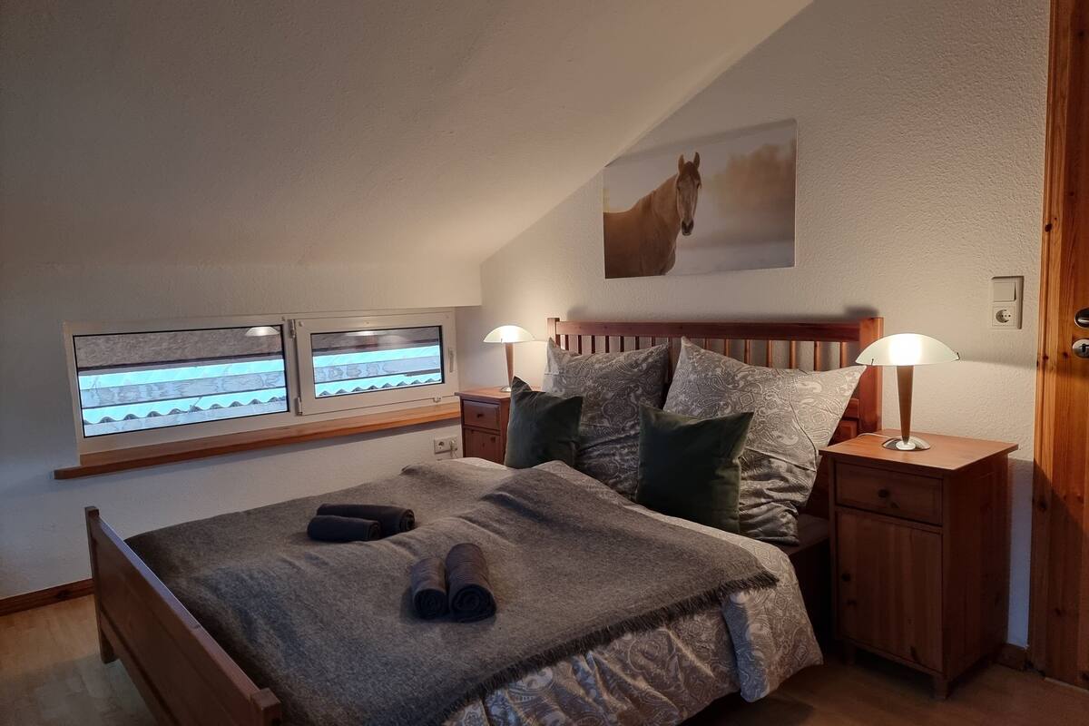 A bedroom with a sloped ceiling, a bed with a patterned bedspread, two bedside lamps, a wooden nightstand, and a mounted TV.