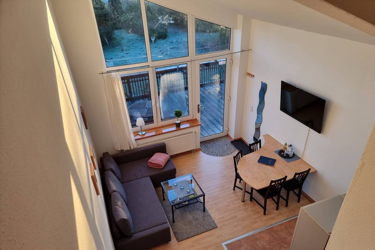 A modern living room with a sofa, coffee table, and dining table. There are large windows and a TV mounted on the wall.