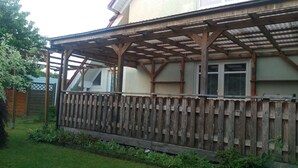 Una pérgola de madera con techo metálico, cerca de madera y una casa con ventanas.