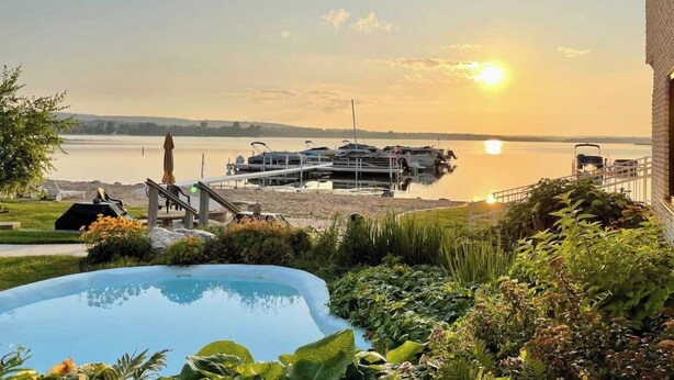 A swimming pool surrounded by lush greenery, a dock with boats, and a view of the sunset over a calm lake.