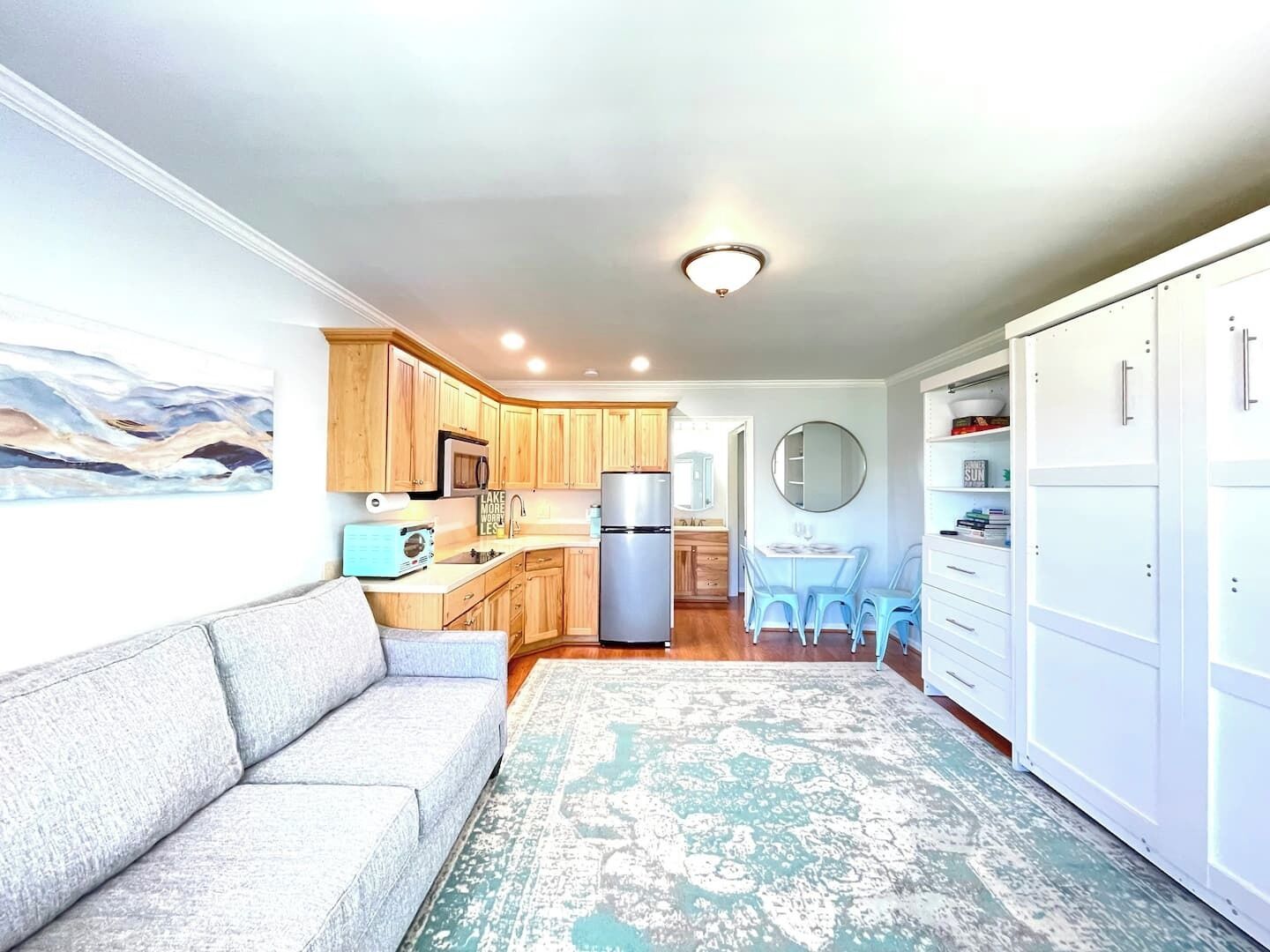 A compact kitchen with wooden cabinets, a microwave, and a refrigerator. There is a dining area with a table and chairs, and a large mirror on the wall.