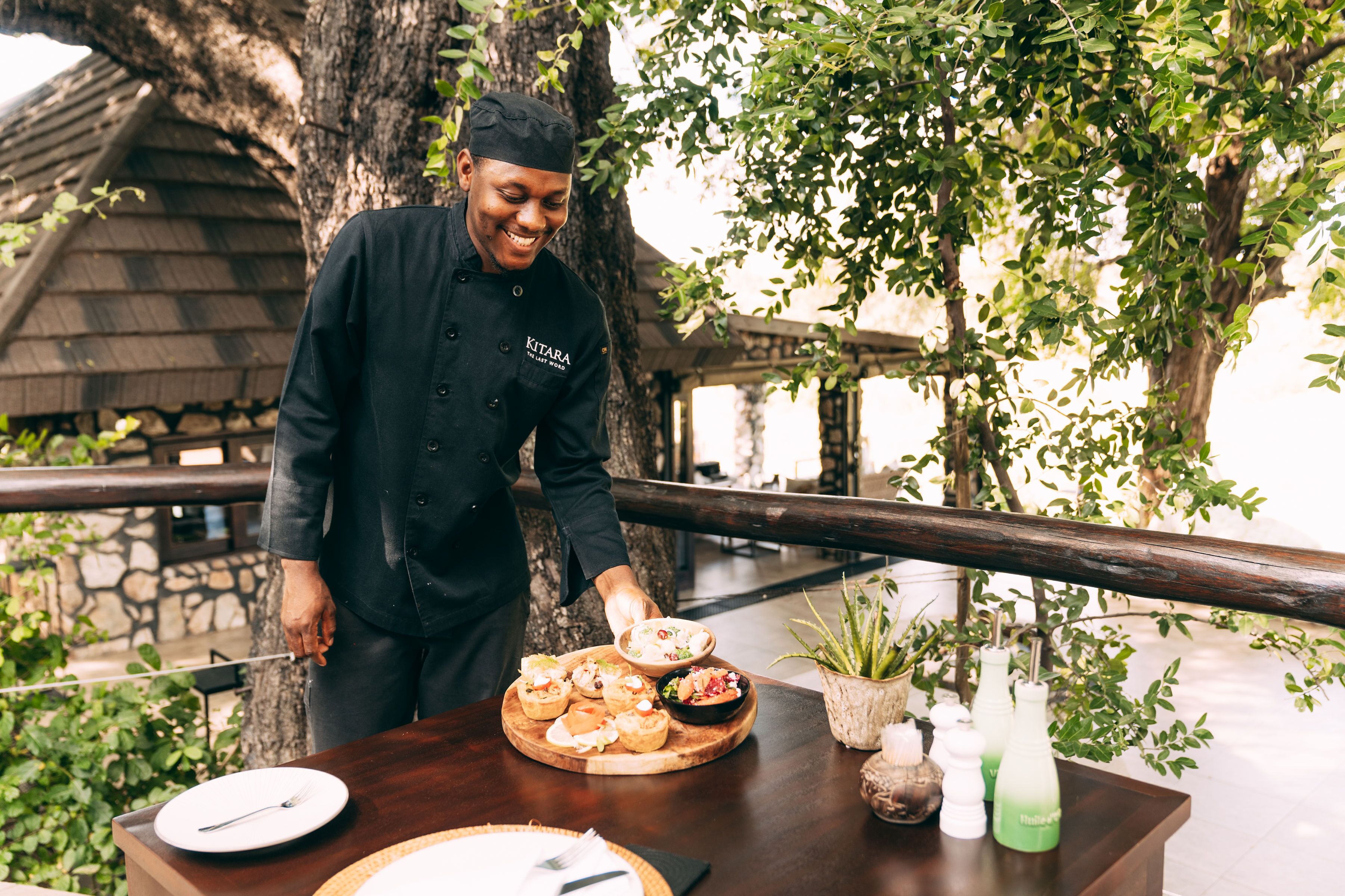 Un chef sirve comida al aire libre, con una mesa de madera, platos y una planta en maceta.