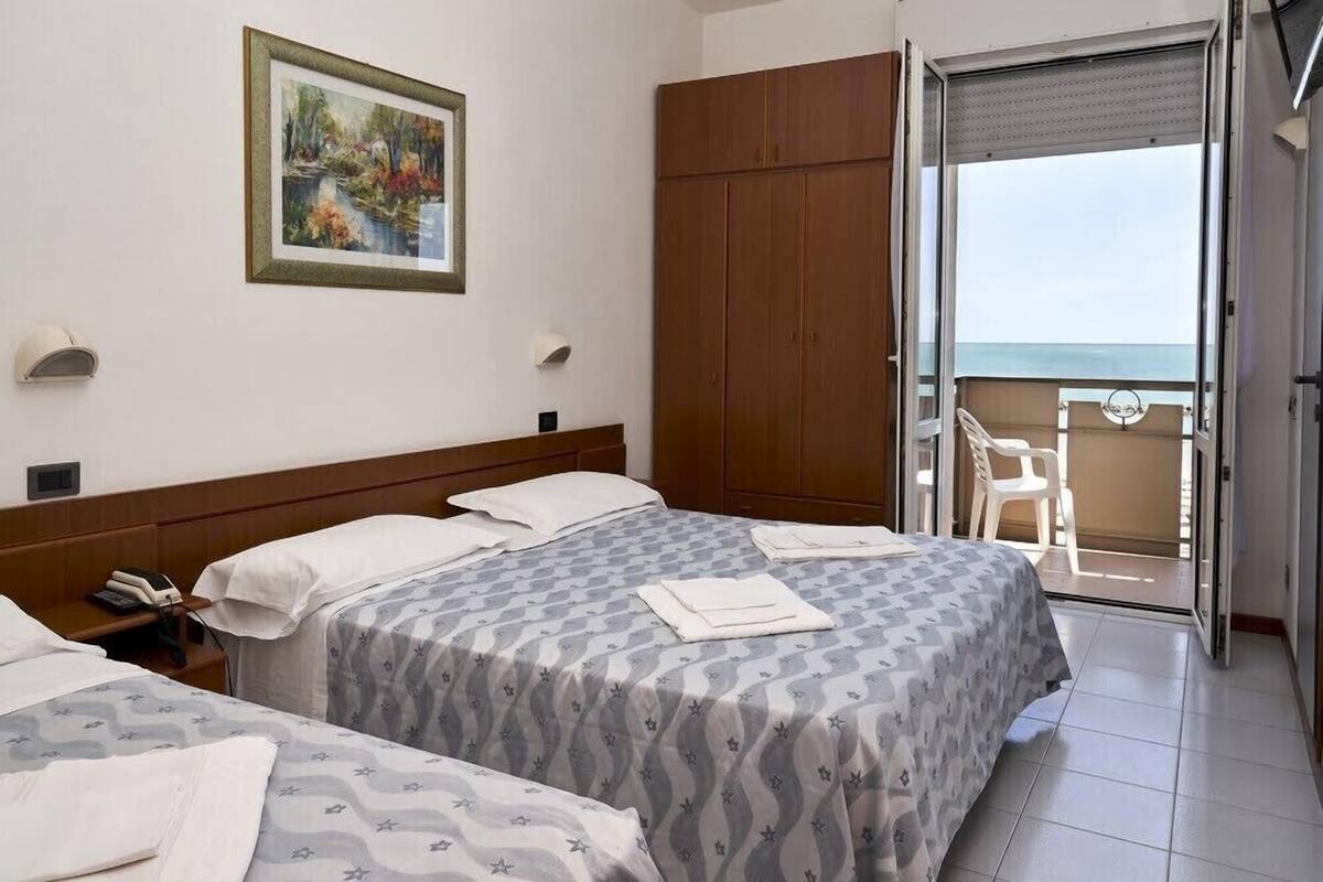 A hotel room with two beds, a painting on the wall, a wardrobe, and a balcony with a view of the sea.