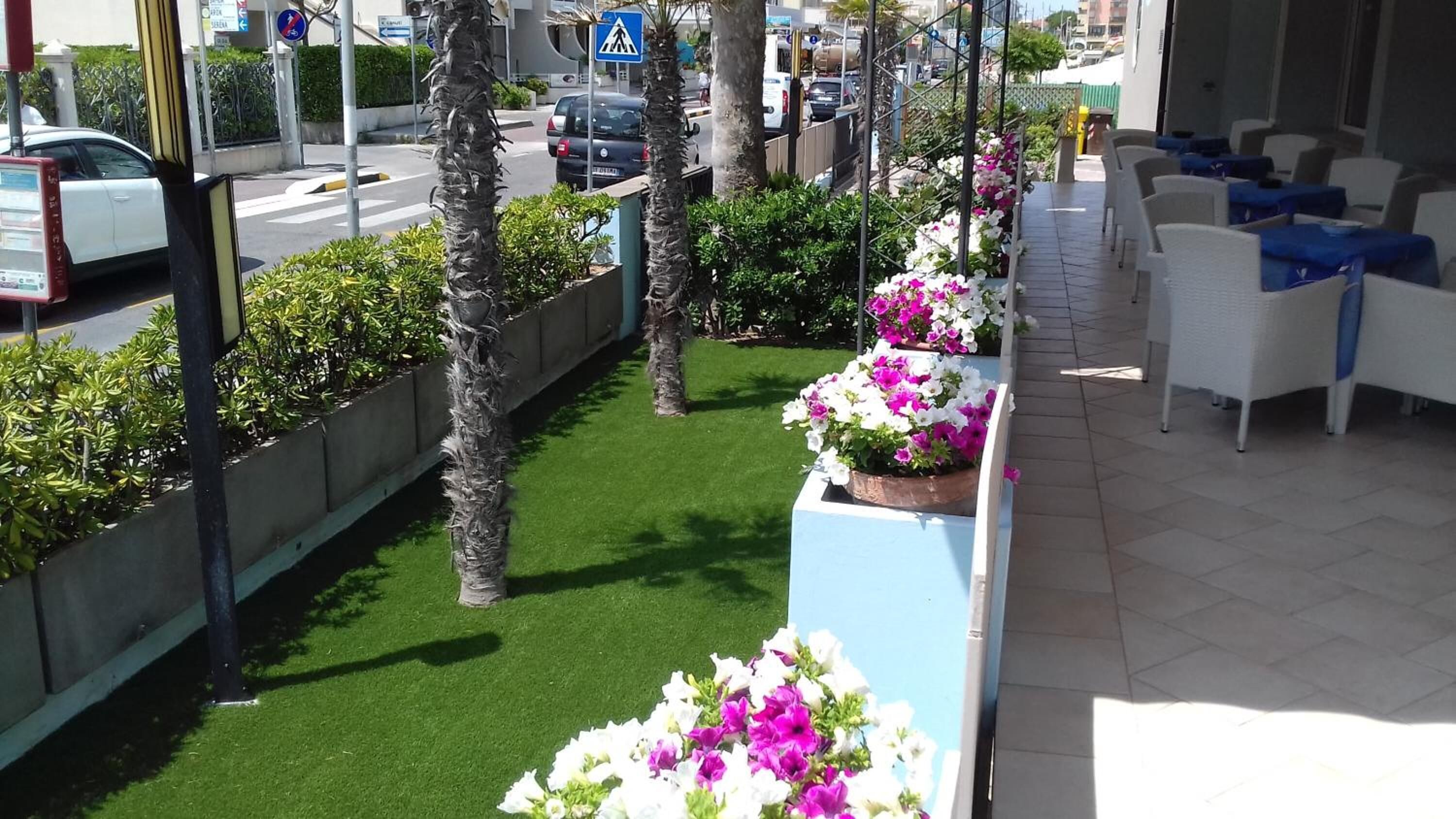 A hotel corridor with indoor plants, a green artificial grass area, and white chairs with blue accents.