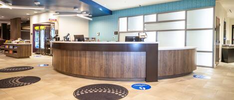 A hotel reception area with a curved reception desk, a coffee machine, and a vending machine. There are two circular mats on the floor, one with a "Thank You" message.