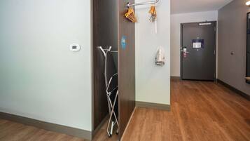 Un pasillo de hotel con piso de madera, un perchero con ganchos, una puerta con un letrero y un termostato en la pared.