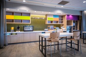A modern cafe with a central island, seating area, and a variety of amenities including a coffee machine, a refrigerator, and a display shelf with items. - Tru by Hilton Minneapolis Mall of America (Bloomington)