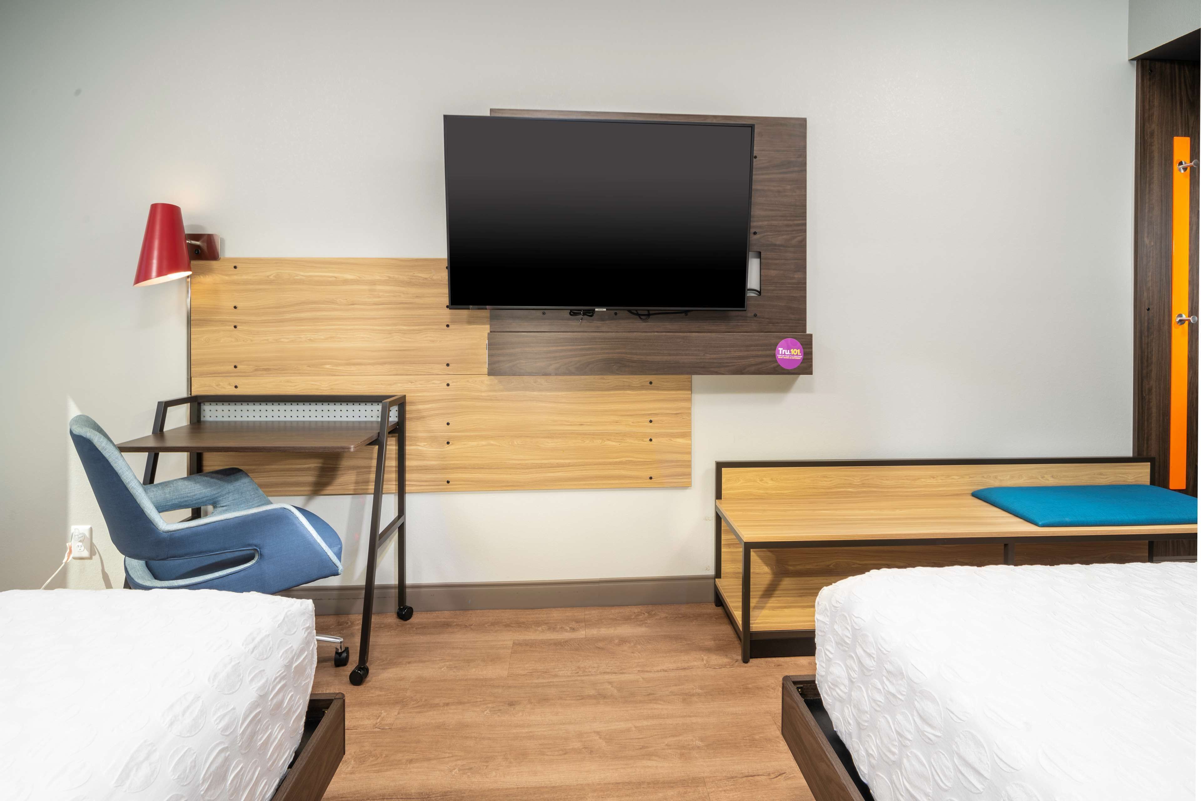 Habitación de hotel con dos camas, un escritorio con silla, un televisor montado en la pared y una mesita con cojín azul.