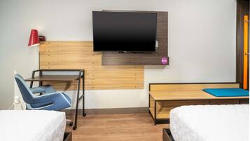 Habitación de hotel con dos camas, un escritorio con silla, un televisor montado en la pared y una mesita con cojín azul.