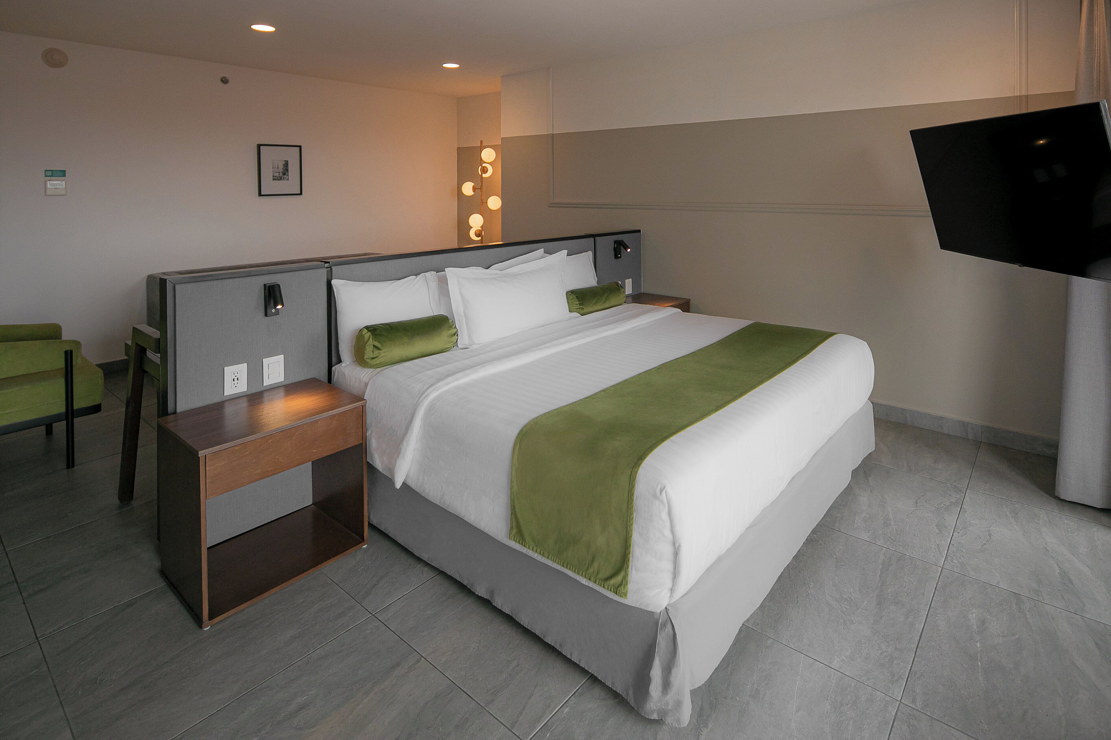 A hotel room with a large bed, a bedside table, a TV, and a green chair.