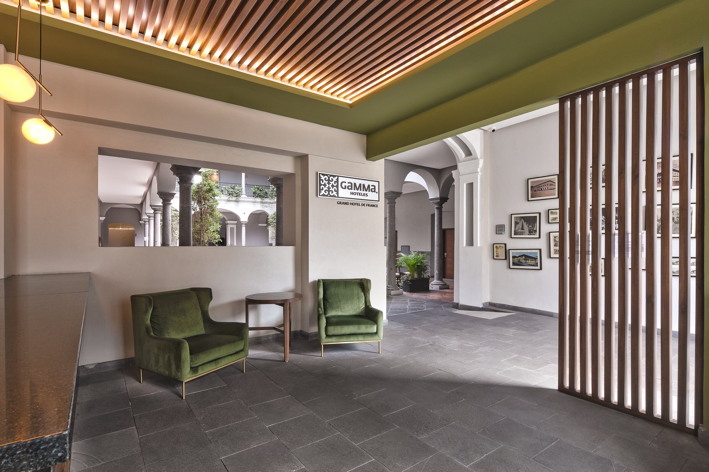Un vestíbulo de hotel con un mostrador de recepción, dos sillones verdes, un separador de madera y cuadros enmarcados en las paredes.