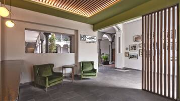 Un vestíbulo de hotel con un mostrador de recepción, dos sillones verdes, un separador de madera y cuadros enmarcados en las paredes.