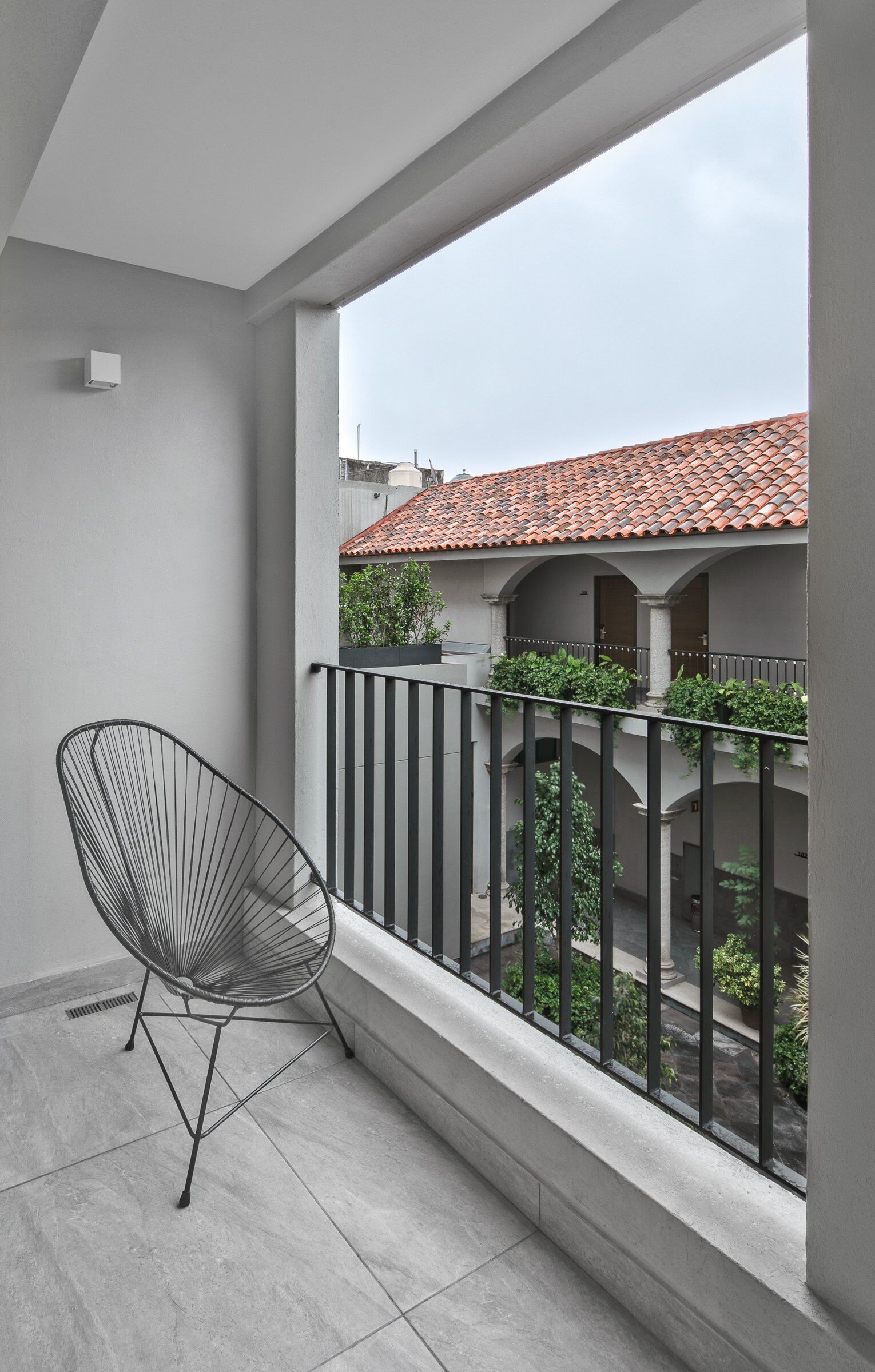 Un balcon avec une chaise en métal, un sol carrelé et une vue sur un immeuble dont le toit est également recouvert de tuiles.