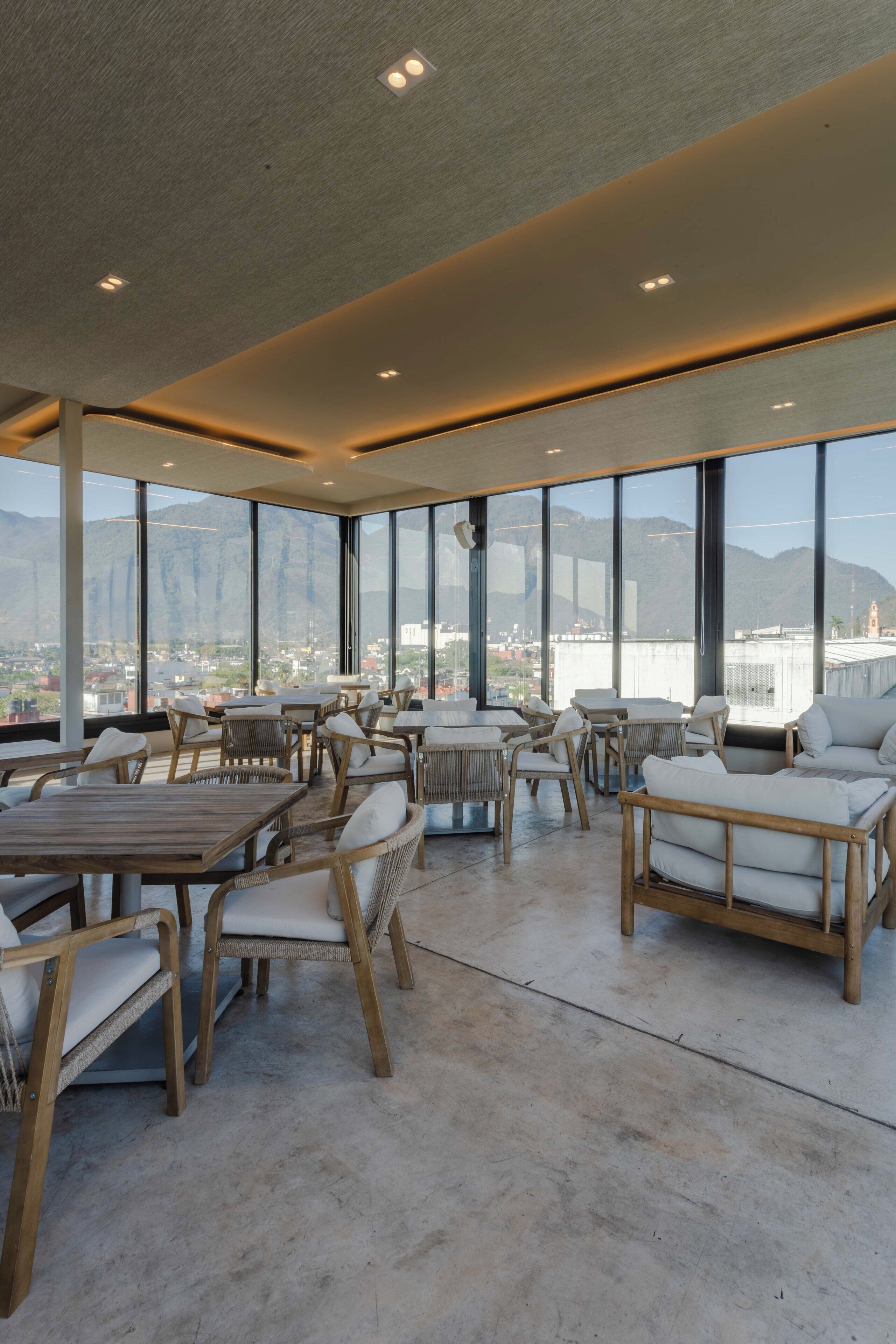 A spacious indoor area with large windows offering a view of mountains and a cityscape. There are several wooden tables and chairs arranged for dining or sitting.