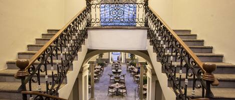 A grand staircase with ornate iron railings leading to a dining area with tables and chairs.