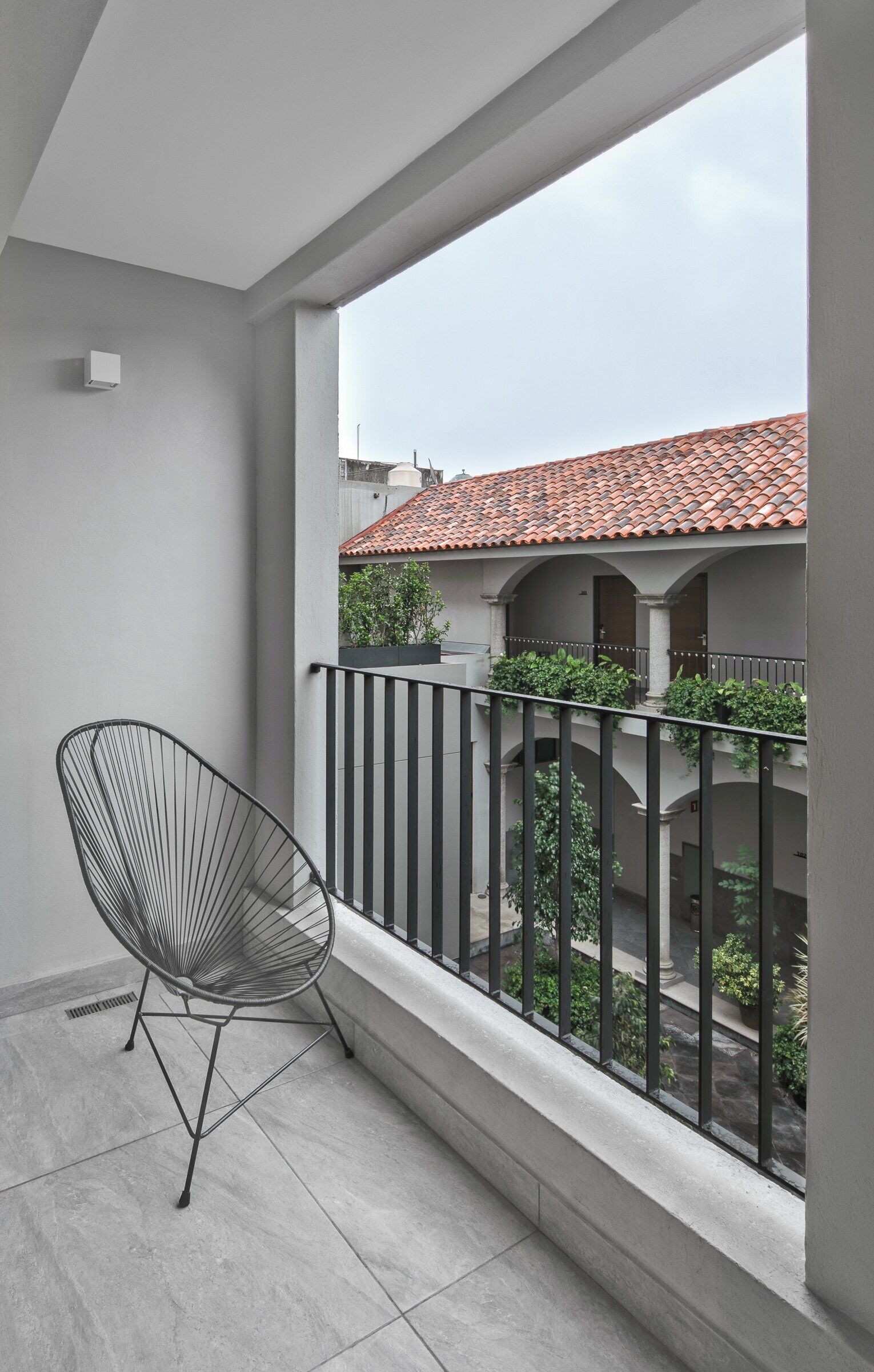 Un balcon avec une chaise en métal, un sol carrelé et une vue sur un immeuble dont le toit est également recouvert de tuiles.