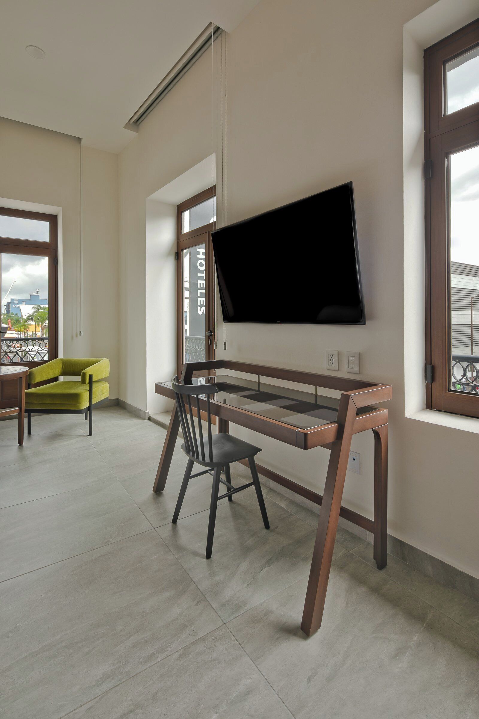 Une chambre d’hôtel moderne avec un bureau en bois, un téléviseur fixé au mur, un fauteuil vert et un balcon offrant une vue sur la ville.