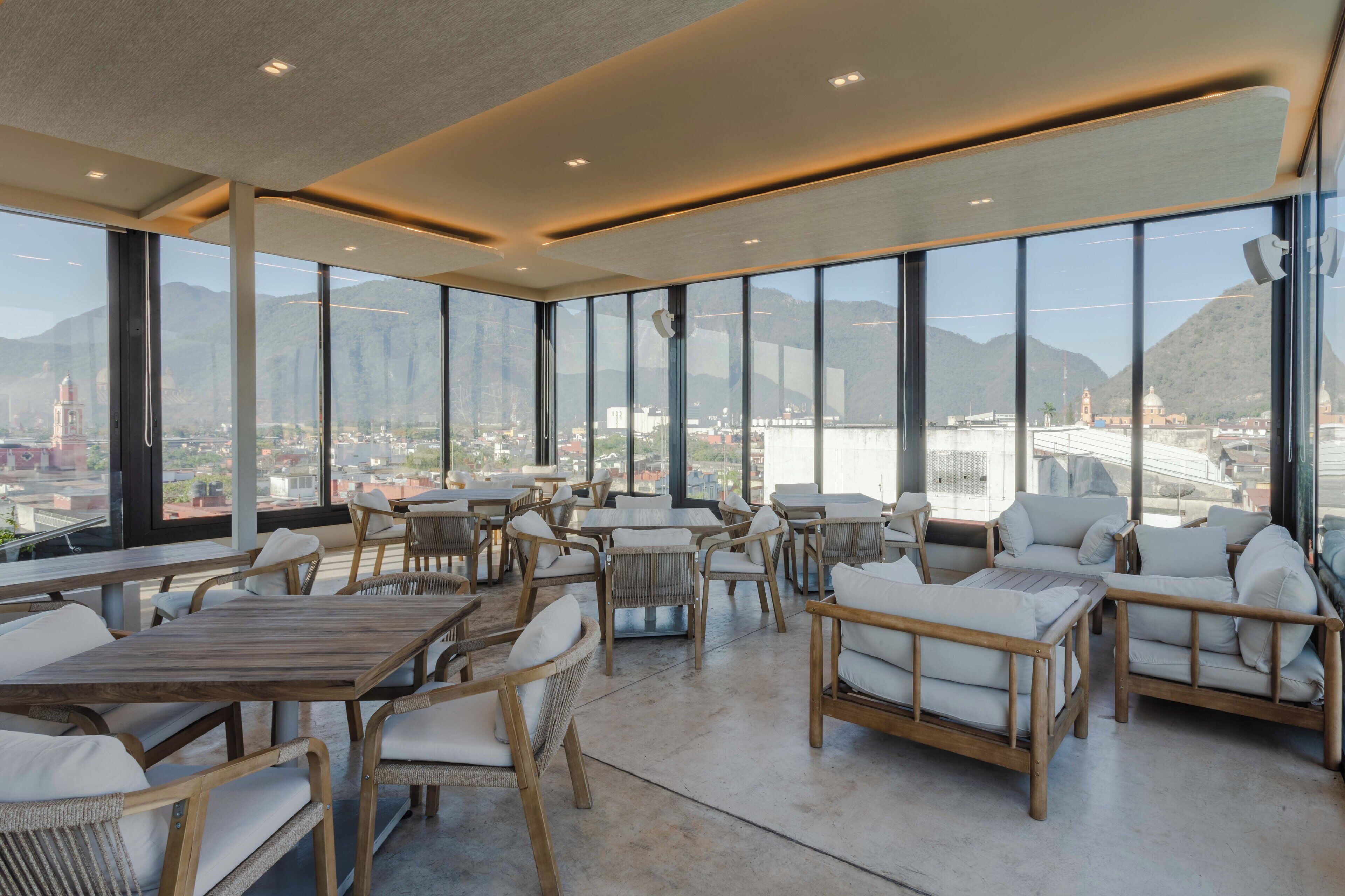 A spacious indoor area with large windows offering a view of a city and mountains. There are wooden tables and wicker chairs with white cushions.
