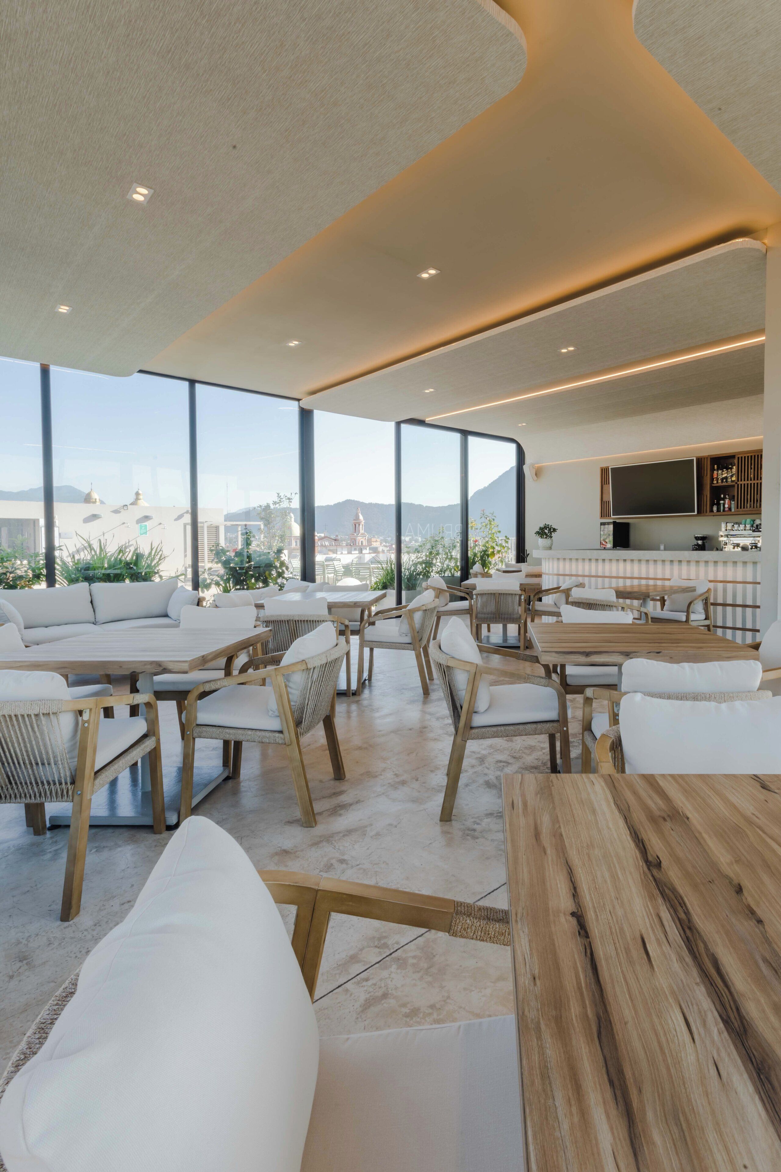 A modern rooftop lounge with wooden furniture, large windows offering city views, and a kitchen area with a flat-screen TV.