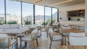 A modern rooftop lounge with wooden furniture, large windows offering city views, and a kitchen area with a flat-screen TV.