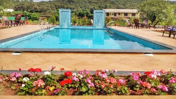 Piscina rectangular con dos pilares de azulejos, rodeada de un parterre con flores rosadas y rojas, y una vista de una colina con árboles y edificios al fondo.