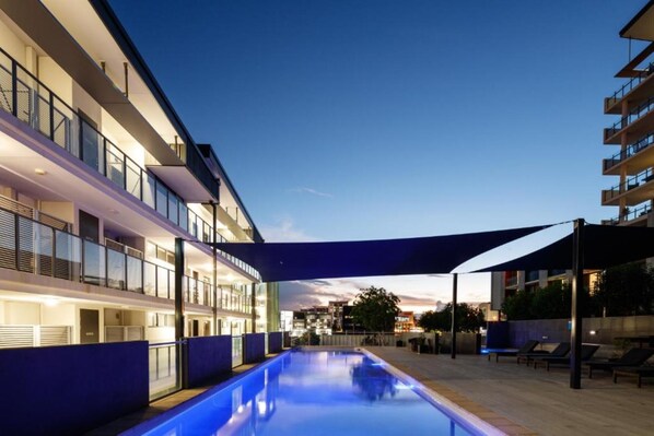 A modern hotel with a swimming pool, outdoor seating, and a view of the city skyline at dusk.