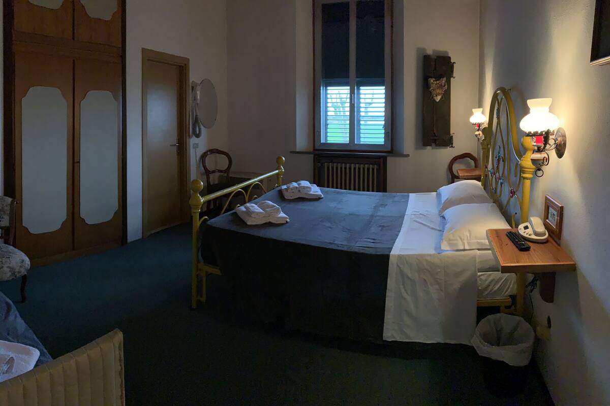 Une chambre d’hôtel avec deux lits, une armoire en bois, une chaise, une petite table avec une lampe et une fenêtre avec des stores.
