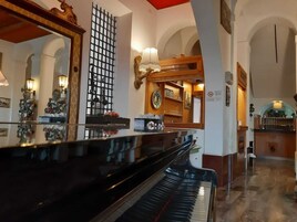 Un piano de cola imponente en un vestíbulo amplio con techo alto, espejos ornamentales y una pared decorativa con un diseño de cuadrícula.