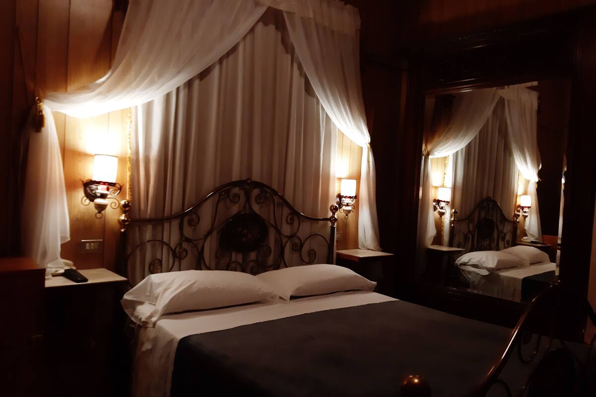 Une pièce faiblement éclairée, avec un lit, une table de chevet, un miroir et une lampe fixée au mur.