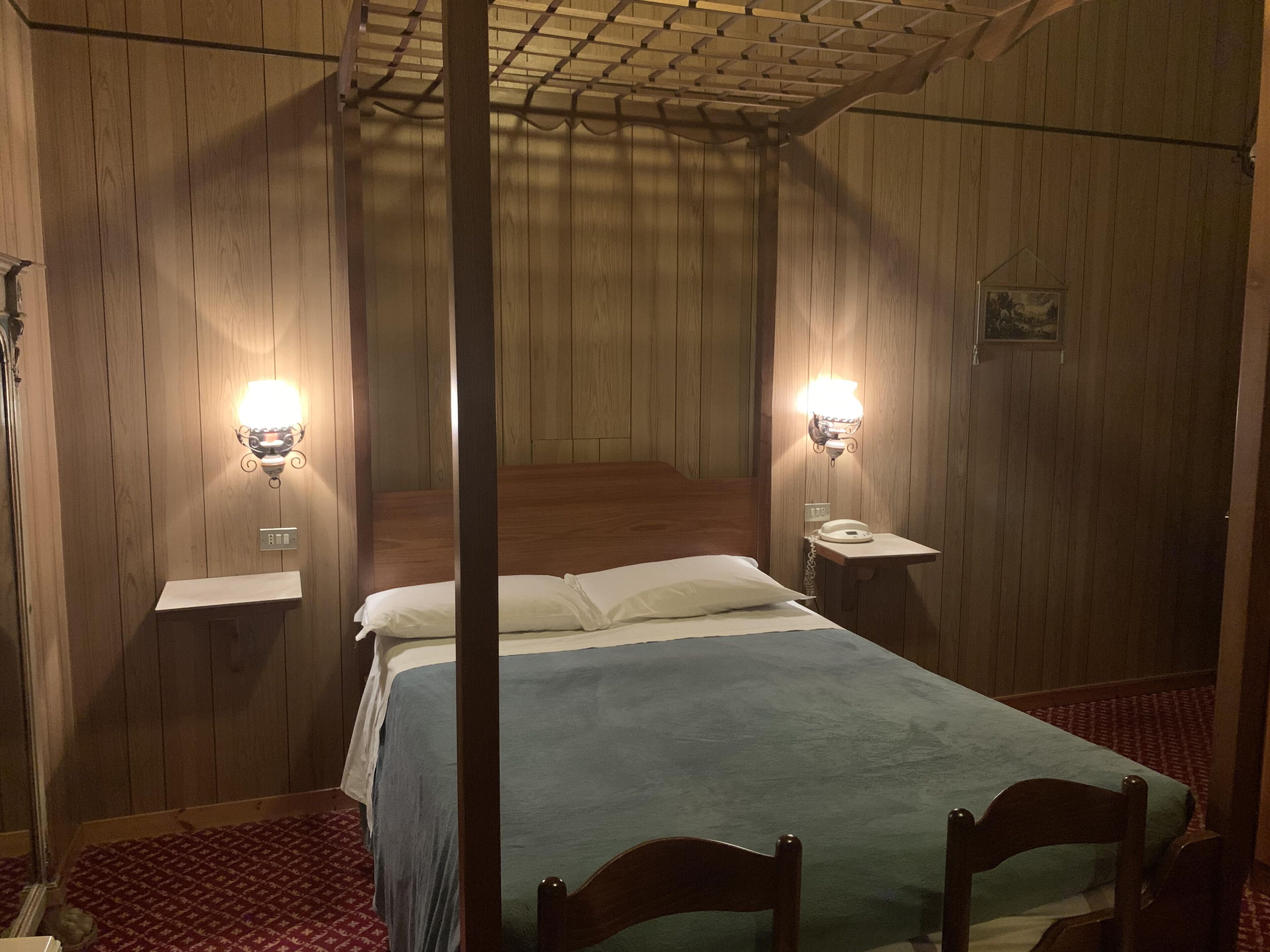 A four-poster bed with a wooden headboard, two bedside tables with lamps, and a red patterned carpet.