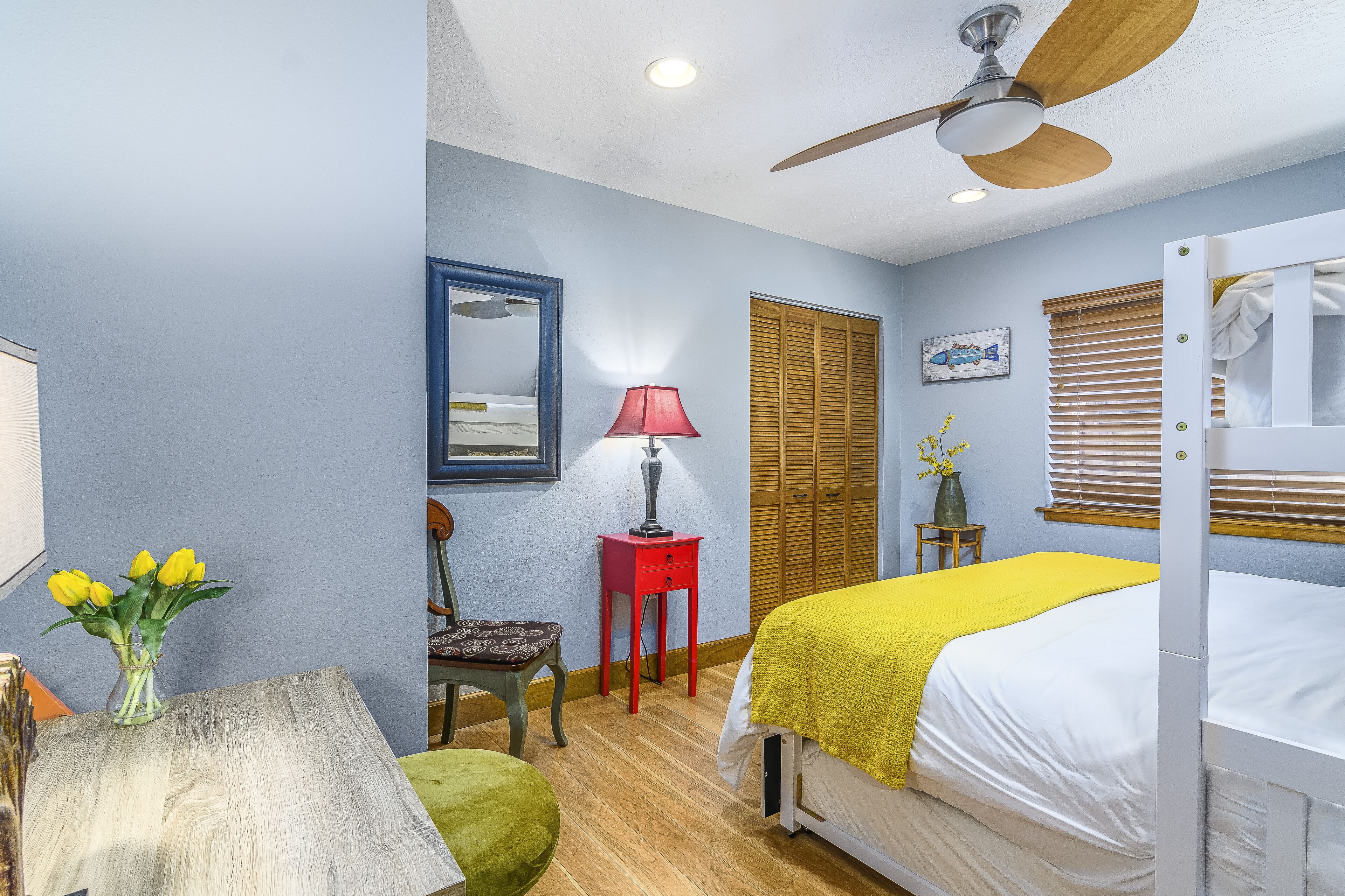 A bedroom with a bed, a desk with a vase of flowers, a chair, a red lamp, and a ceiling fan.