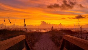 Ein Holzsteg führt zu einem Strand, im Hintergrund ist ein Sonnenuntergang zu sehen.