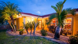 Ein Haus mit rotem Ziegeldach, einem gepflegten Garten mit Palmen und Topfpflanzen sowie einer sichtbaren Tür zum Innenbereich.