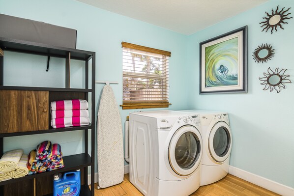 A laundry area with a washing machine, shelves with folded towels, and a window with blinds. - Family-Friendly Home: Heated Pool & Spa: 0.3 Mi Walk to Beach Watch the Rockets! (Cocoa Beach)