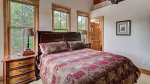 A bedroom with a large bed, wooden headboard, and multiple windows. There is a dresser with drawers and a lamp beside the bed. - The Luxurious Lincoln Hot Tub (Breckenridge)
