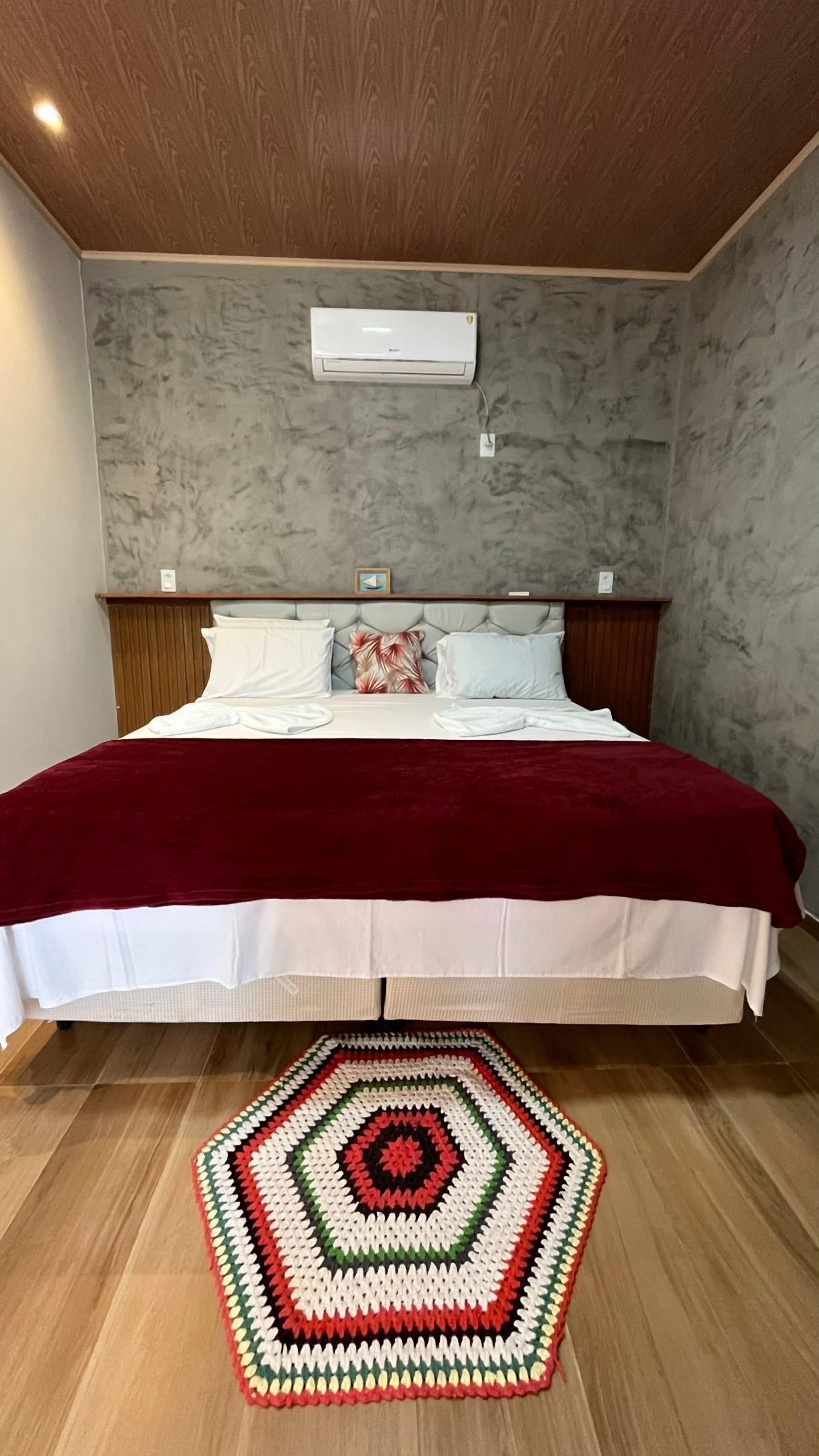 A bedroom with a bed, a red blanket, a geometric rug, and an air conditioning unit.
