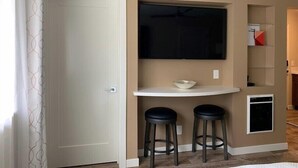 A room with a white door, a white console table with a bowl, two black stools, a flat-screen TV mounted above the table, and a small appliance on the wall.