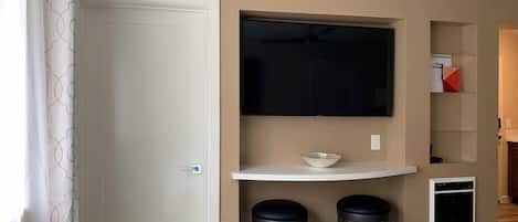 A room with a white door, a white console table with a bowl, two black stools, a flat-screen TV mounted above the table, and a small appliance on the wall.