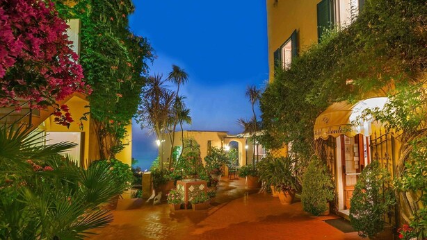 A courtyard with a pathway, potted plants, and a building with a yellow facade and green shutters. - Hotel Monti (Casamicciola Terme)
