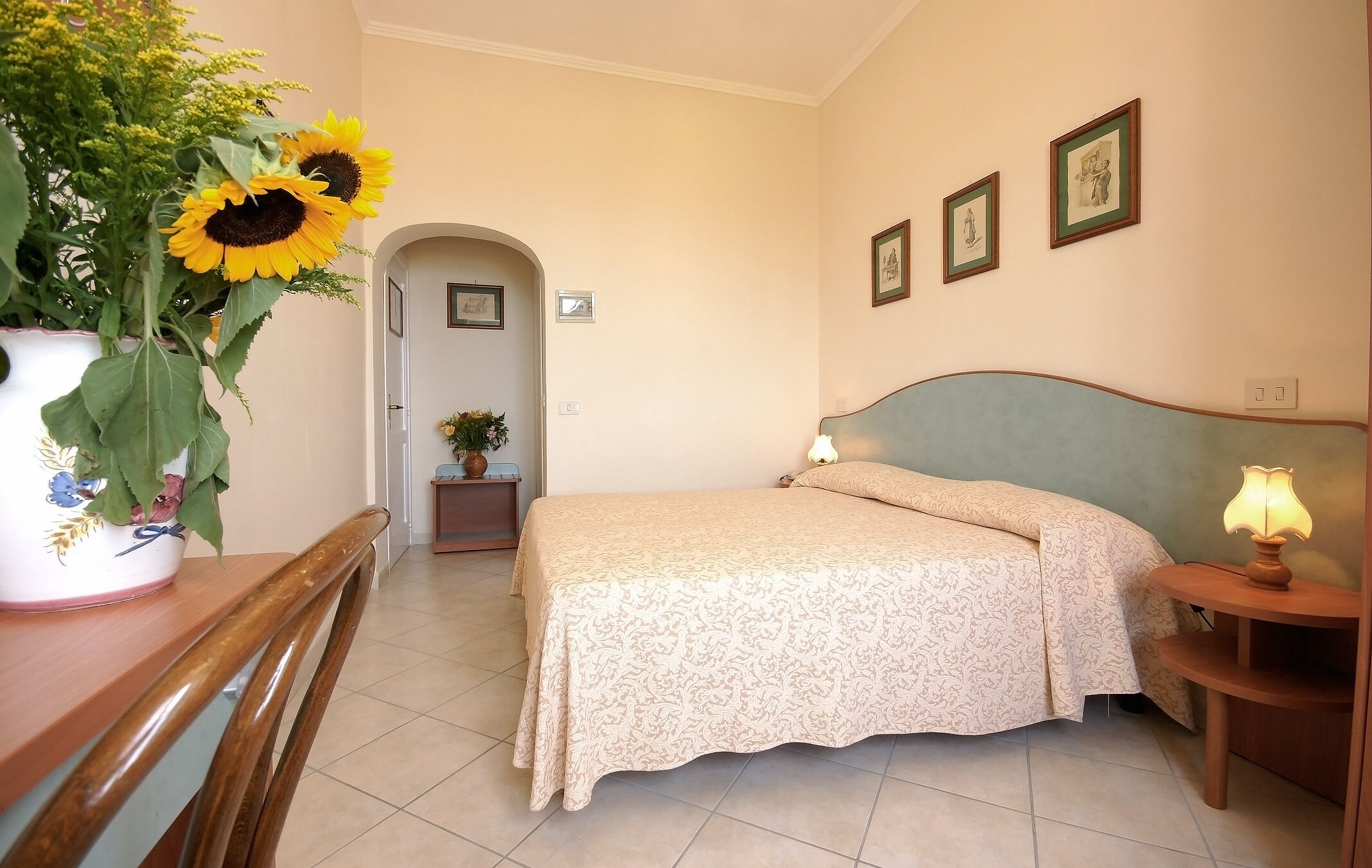 A bedroom with a bed, a bedside table with a lamp, a wooden chair, a vase with sunflowers, and a door leading to another room.