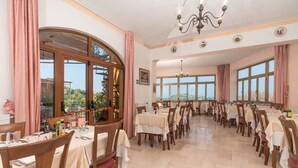 A dining area with multiple tables and chairs, large windows, and a chandelier. - Hotel Monti (Casamicciola Terme)