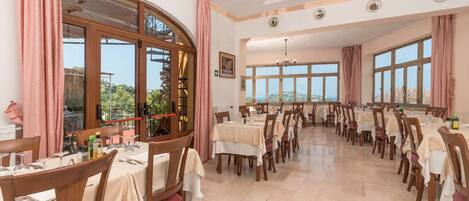 Un comedor con varias mesas y sillas, ventanas amplias y un candelabro.