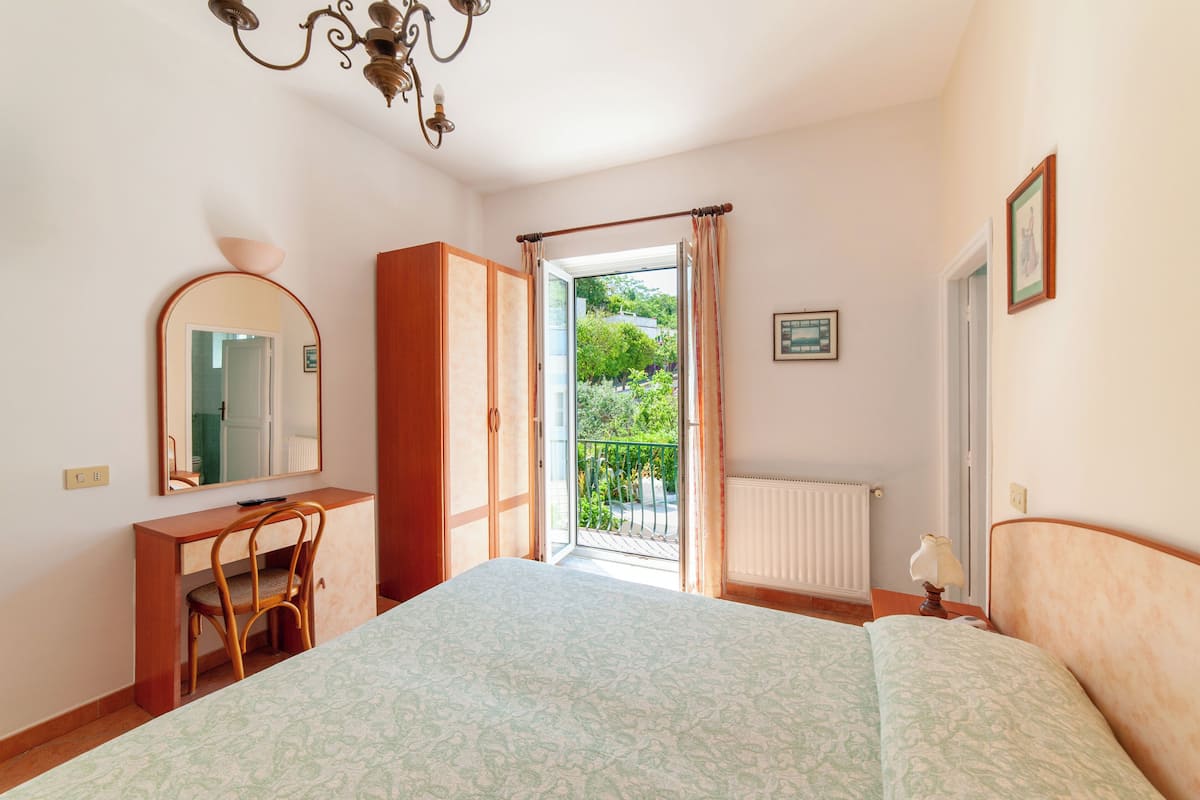 Een slaapkamer met een bed, een bureau, een stoel, een kledingkast, een spiegel, een raam met gordijnen en een radiator.
