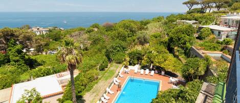 A rooftop pool with lounge chairs, surrounded by greenery and overlooking a coastal view.