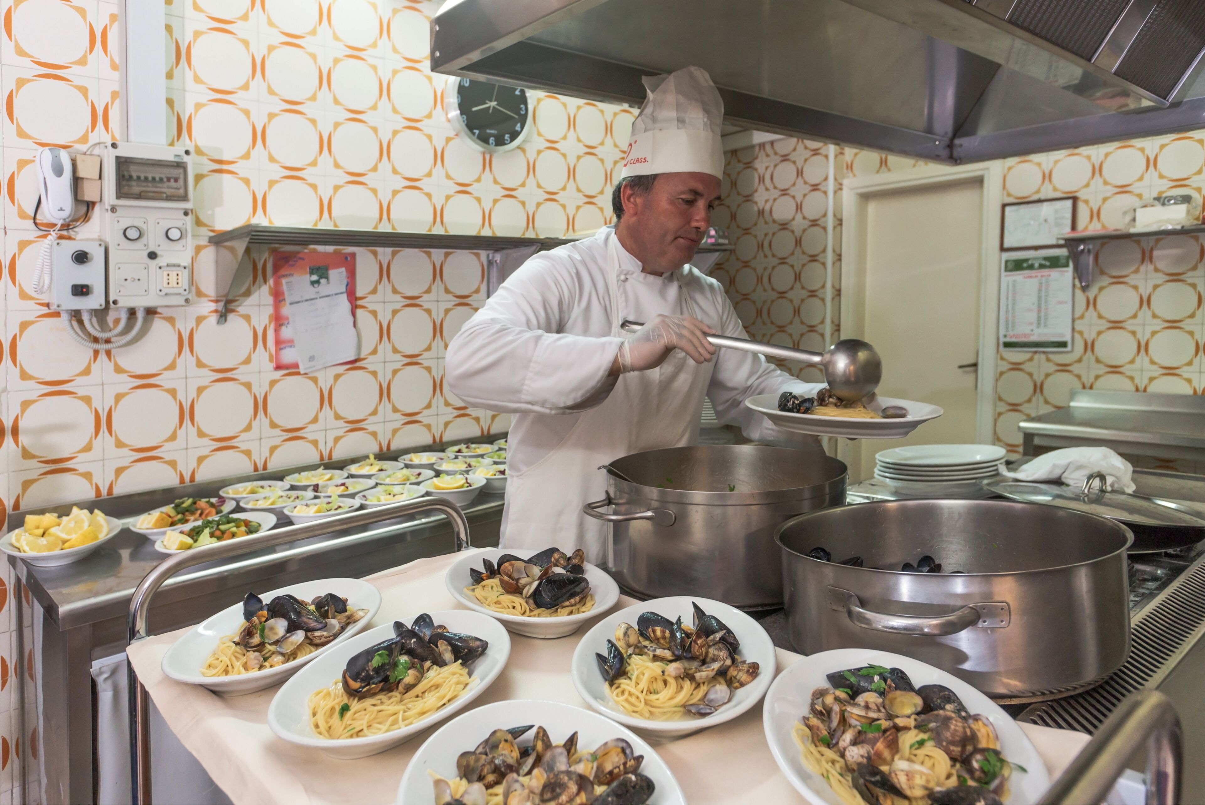 A chef in a kitchen with multiple cooking stations, stainless steel pots, and plates of pasta.