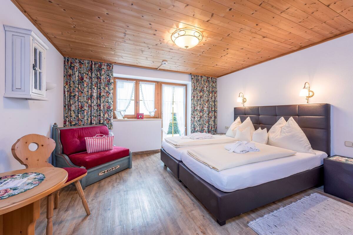 A bedroom with a wooden ceiling, a bed with white bedding, a red sofa, and a small table with a floral tablecloth.
